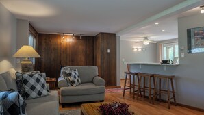 A living room with a sofa, a coffee table, a dining area with bar stools, wooden paneling, and recessed lighting.