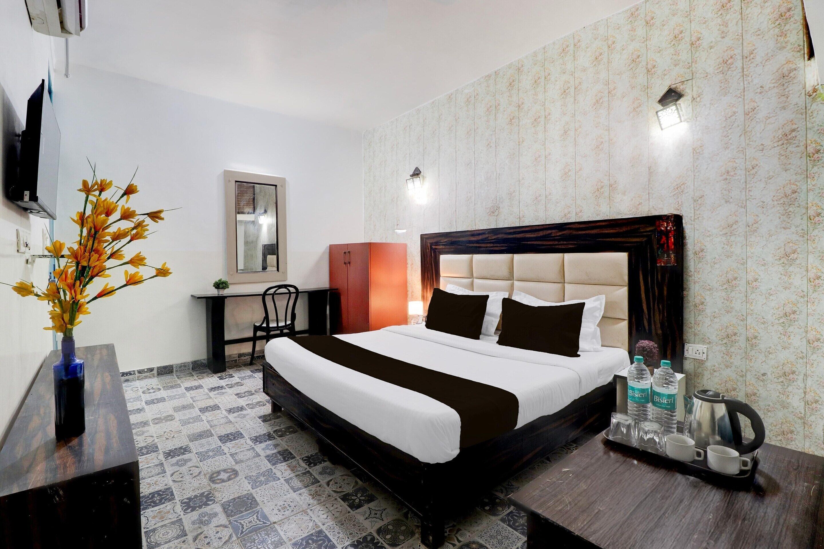 A hotel room with a bed, a desk, a chair, a TV, a vase with flowers, and a tray with water bottles and cups.