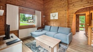 L’intérieur d’un chalet en bois est aménagé avec un canapé d’angle, une petite table, un téléviseur et une fenêtre donnant sur un paysage verdoyant.