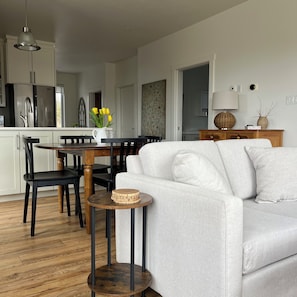 Una sala de estar moderna con un sofá, una mesa de comedor con sillas, una pequeña mesa auxiliar y una cocina al fondo.