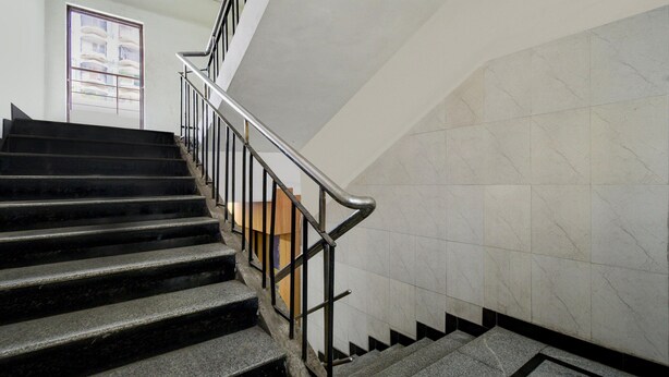 Escada com corrimão de metal, degraus pretos e paredes revestidas de azulejos brancos.