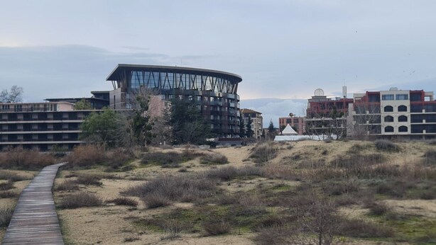 Un edificio moderno con facciata in vetro curvo, una passerella in legno ed edifici bassi circostanti.