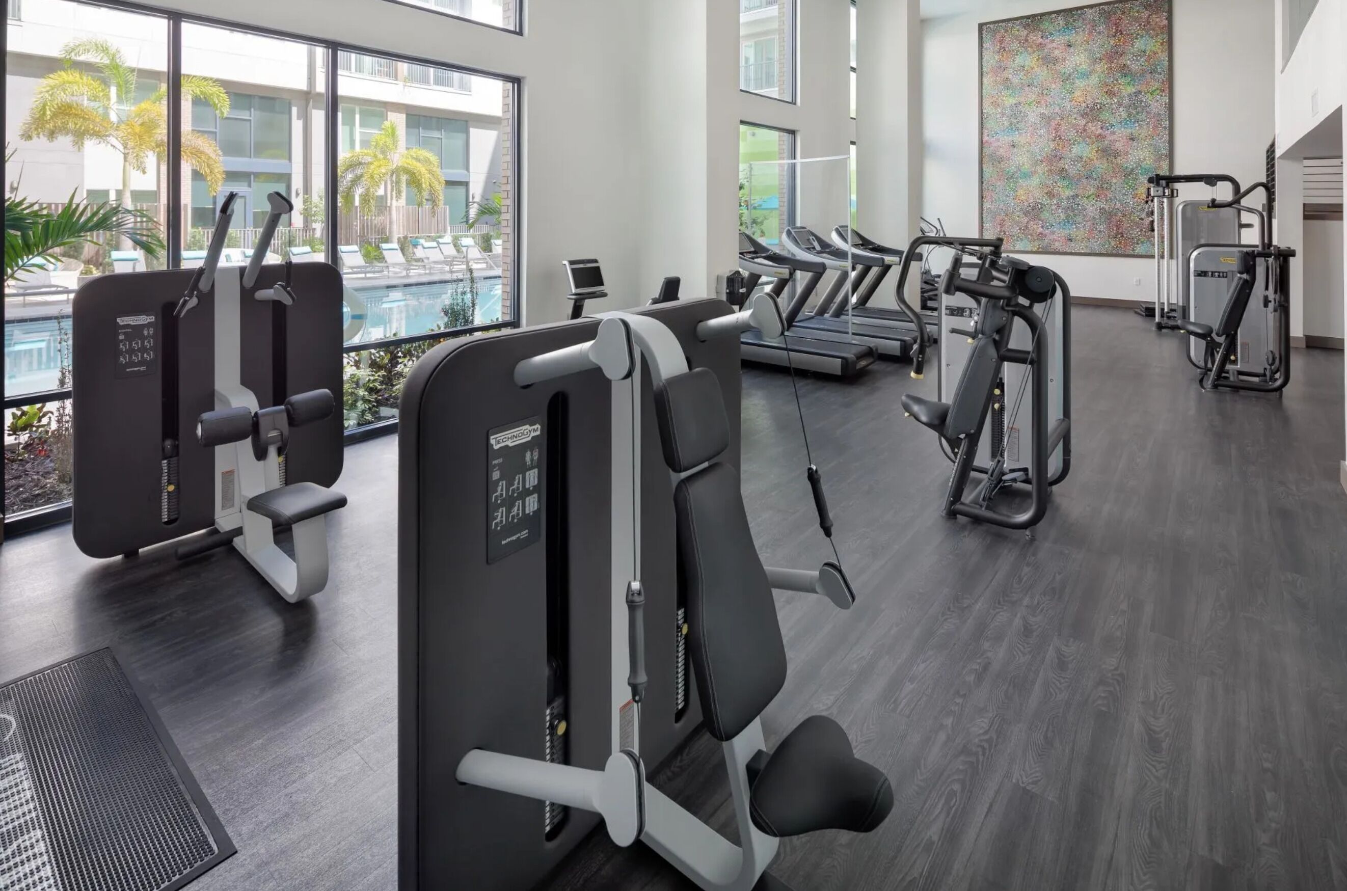 A modern gym with cardio machines, weight training equipment, and a view of a pool and palm trees.