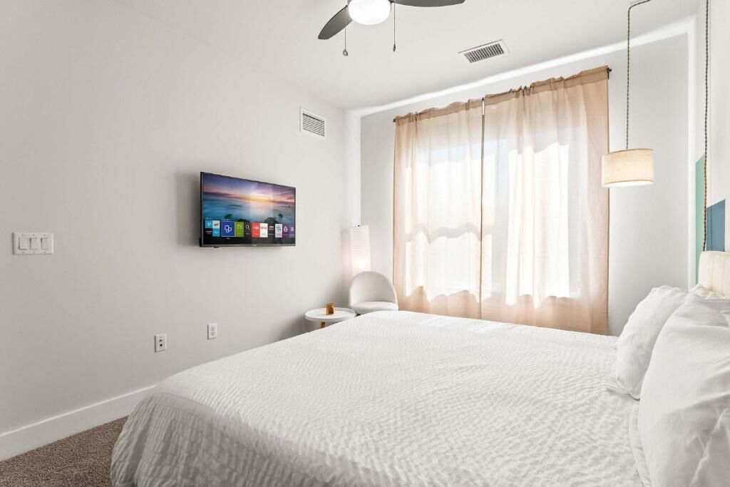 A hotel room with a bed, a TV mounted on the wall, a ceiling fan, and a window with curtains.