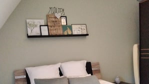 Quarto com cama, mesa de cabeceira, cômoda e prateleira na parede com diversos objetos.