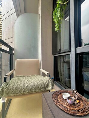 Ein Balkon mit gepolstertem Stuhl, einem gewebten Tablett mit Dekoration und einer hängenden Pflanze.