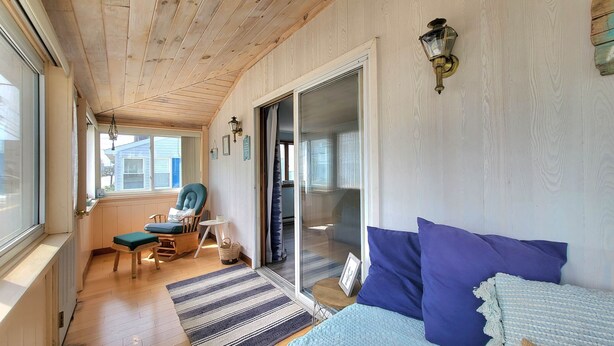 A room with a wooden ceiling, a sliding glass door, a blue sofa, a chair with a cushion, a small table, and a rug. - Sunset Sanctuary by Wells Beach (Wells)