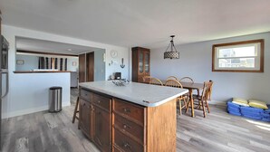 A kitchen with a central island, wooden cabinets, a dining area with chairs, a chandelier, and a window with a view. - Sunset Sanctuary by Wells Beach (Wells)