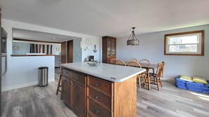 A kitchen with a central island, wooden cabinets, a dining area with chairs, a chandelier, and a window with a view.
