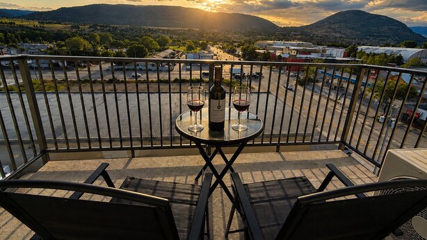 A balcony with two chairs, a table, and two wine glasses with bottles, overlooking a parking lot and a hilly landscape during sunset. - Top Floor Walk to Beach & Casino (Penticton, BC)