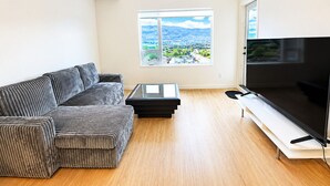 A modern living room with a sectional sofa, a flat-screen TV, a coffee table, and a large window offering a city view. - Top Floor Walk to Beach & Casino (Penticton, BC)