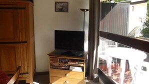 A room with a wooden cabinet, a television on a stand, and a window with a view of a building and greenery. - Ski-in/ski-out studio with balcony (Enchastrayes)