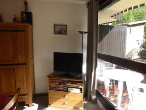 A room with a wooden cabinet, a television on a stand, and a window with a view of a building and greenery. - Ski-in/ski-out studio with balcony (Enchastrayes)