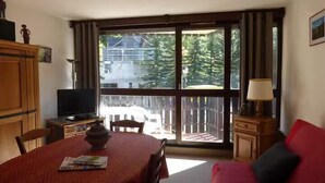 A room with a dining table, a television, a sofa, and a large window with a view of trees and buildings. - Ski-in/ski-out studio with balcony (Enchastrayes)