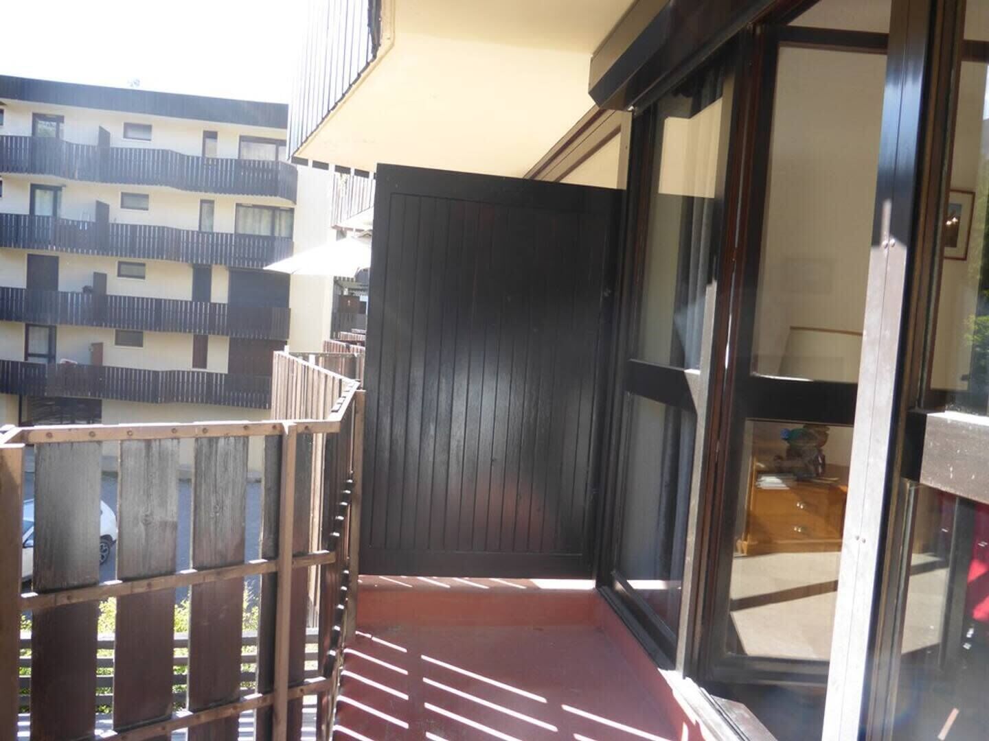 A balcony with a wooden railing, a red floor, and a view of adjacent buildings.