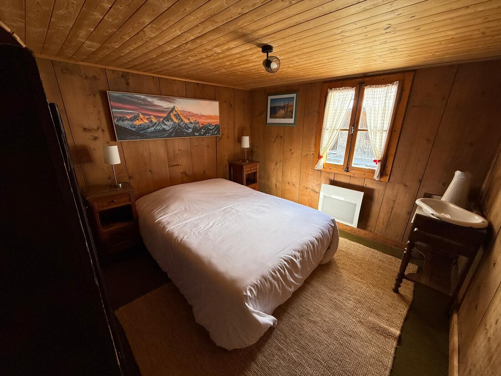 Una habitación de madera con cama, una ventana con cortinas, un radiador y un cuadro en la pared.