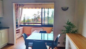 Ein Zimmer mit Esstisch, Stühlen, Küchenzeile, einem Fenster mit Blick auf Palmen und einer Zimmerpflanze.