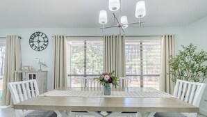Un área de comedor con una mesa de madera, sillas blancas, un jarrón de flores, un reloj de pared y una ventana con cortinas.
