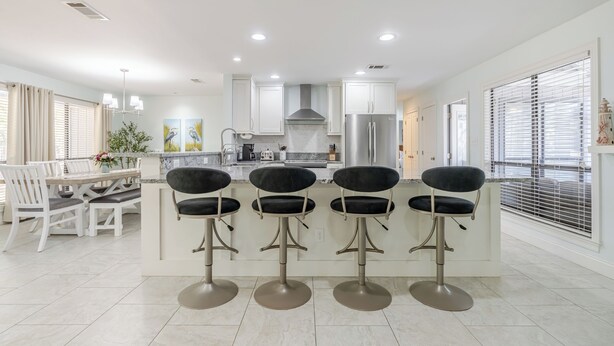 Una cocina moderna con una isla central, taburetes de bar, un comedor con mesa y sillas blancas, y un ventanal con persianas.