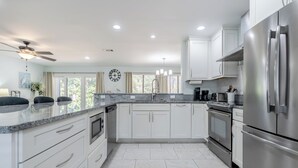 Una cocina moderna con armarios blancos, electrodomésticos de acero inoxidable, encimera de mármol y un amplio ventanal con cortinas.