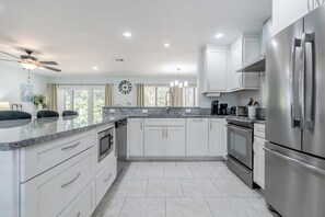 A modern kitchen with white cabinets, stainless steel appliances, a marble countertop, and a large window with curtains. - Sandestin 3BDR- Sleeps 8/Golf Cart/Pet /Pool/Beach (Miramar Beach)