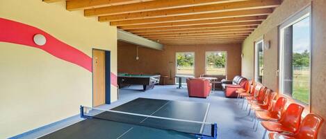 A ping pong table, seating area, and a billiards table in a covered indoor space with wooden beams and large windows.