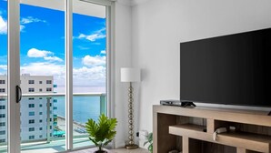 A modern living room with a large TV, a wooden coffee table, a floor lamp, and a view of the ocean through floor-to-ceiling windows. - Stunning Beachfront Penthouse - Panoramic Ocean Views - 1 Bed / 1 Bath -Paradise (Hollywood)
