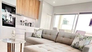 Een moderne slaapkamer met een groot bed, een glazen salontafel en een keukenruimte zichtbaar door een geopende deur.