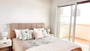 Een slaapkamer met een groot bed, nachtkastje, lamp en een glazen schuifdeur naar een balkon.