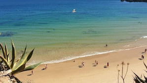 Een strand met helder water, mensen die zonnebaden en een rotsachtige landtong in de verte.