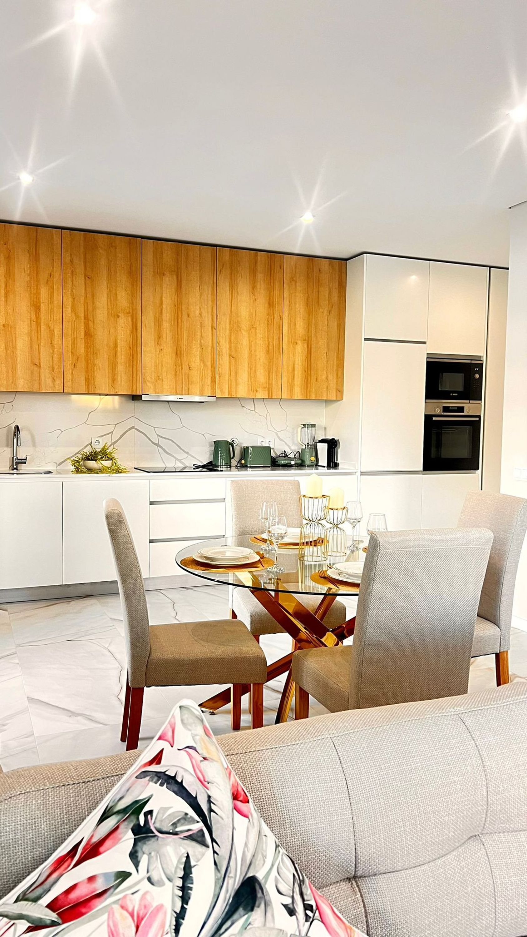 Eine moderne Küche mit Holzausstattung, eine Essecke mit Glastisch und ein Sofa mit einem Blumenkissen.