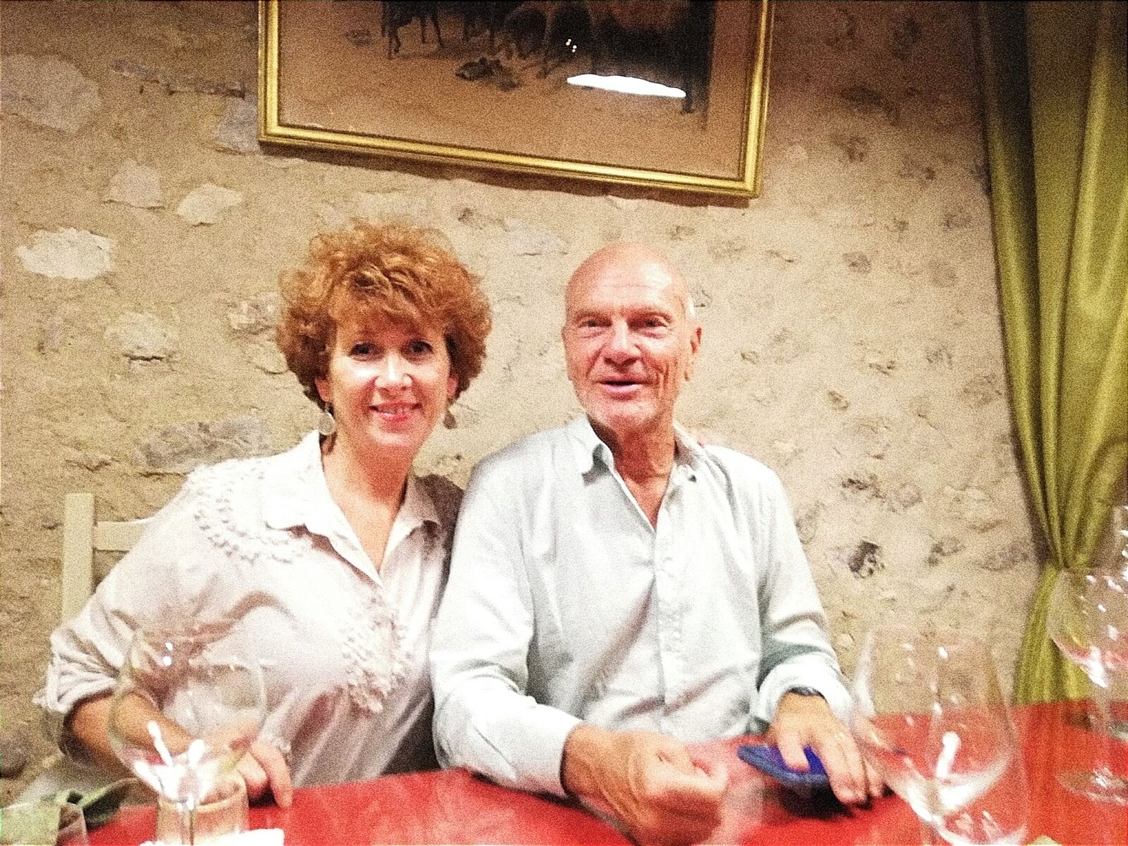 Two people seated at a table with a red tablecloth, a framed picture on the wall, and a green curtain.