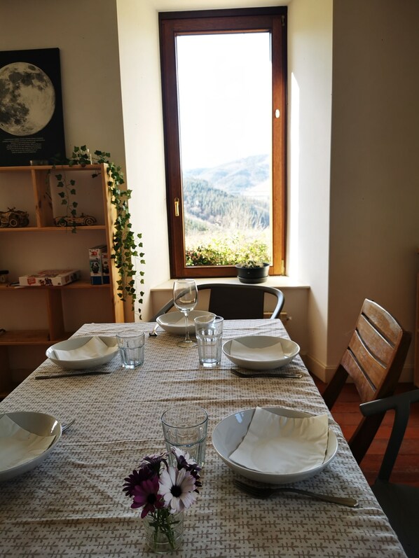 Une table à manger dressée avec des assiettes, des verres et un vase de fleurs, donnant sur une fenêtre qui offre une vue sur une forêt et des montagnes.