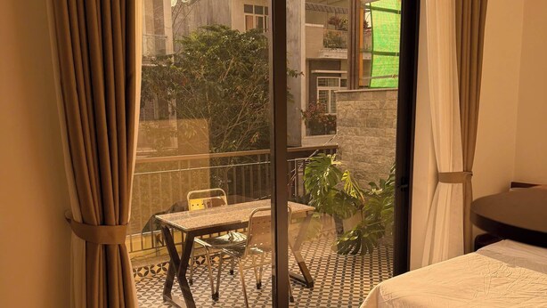 A hotel room with a bed, a dining table with chairs, a balcony with a view of buildings and greenery, and a sliding glass door. - THIÊN PHÚC HOTEL (Nghia Lo)