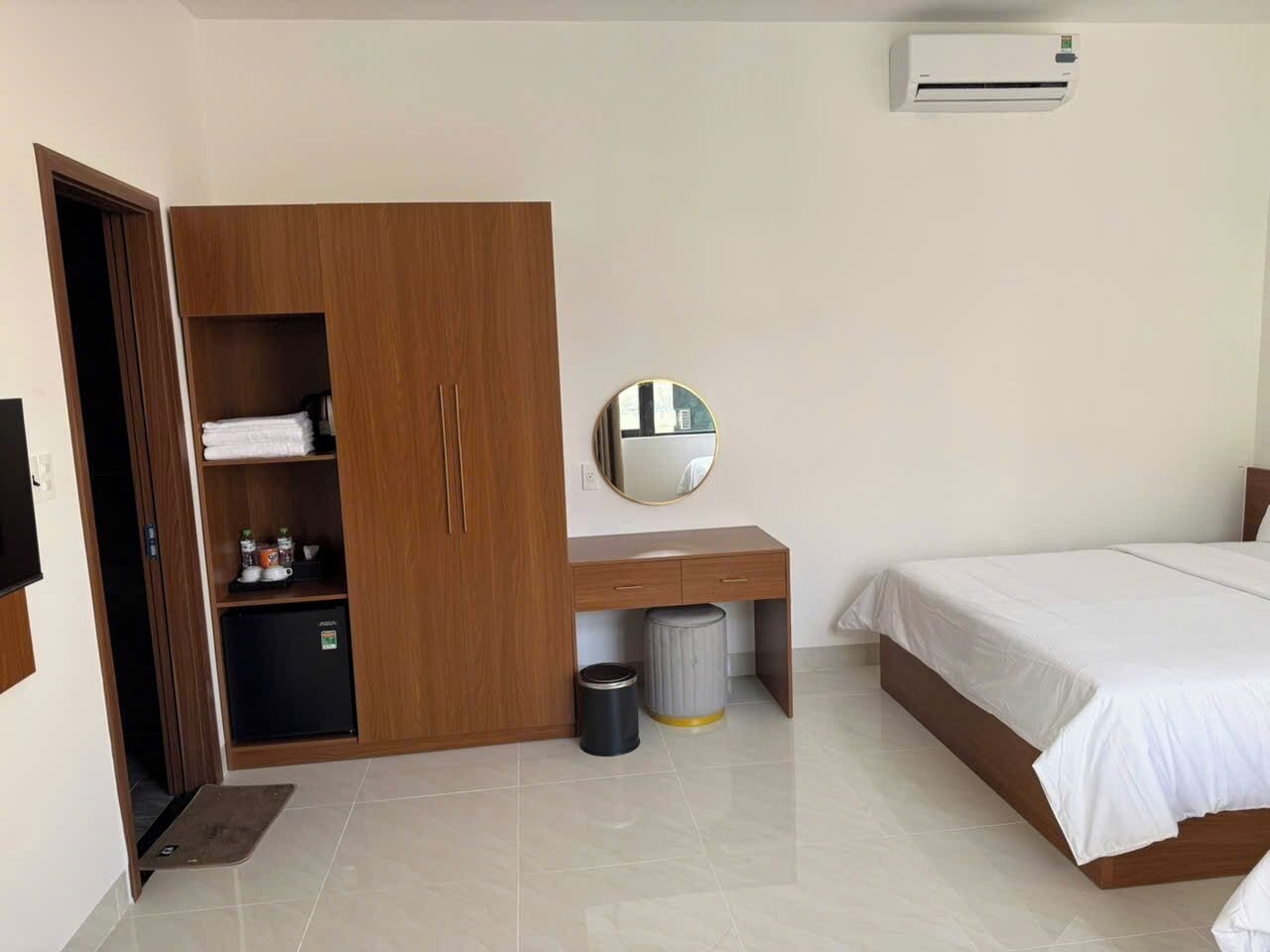 A bedroom with a bed, a wooden wardrobe, a desk with a mirror, and an air conditioning unit.