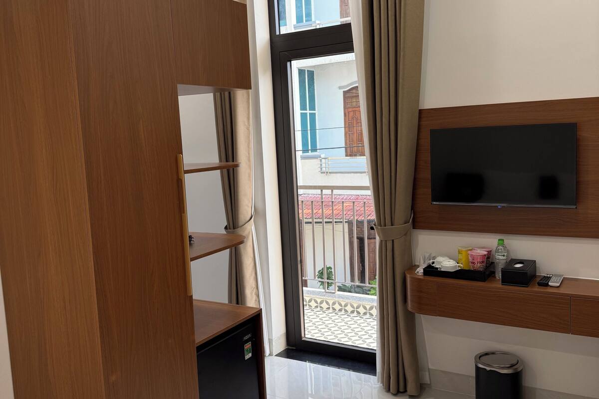 Una habitación moderna con un armario de madera, un televisor montado en la pared, un escritorio pequeño con un cesto de basura y una ventana con cortinas.