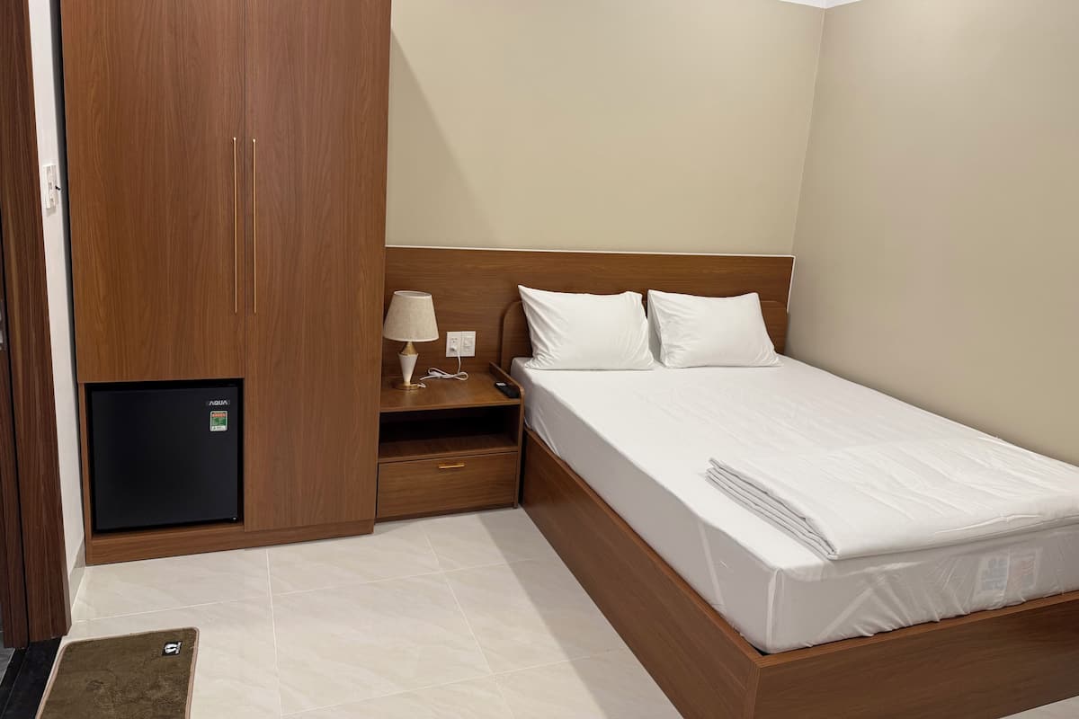 Una habitación de hotel con estructura de cama de madera, mesita de noche con lámpara y minibar.