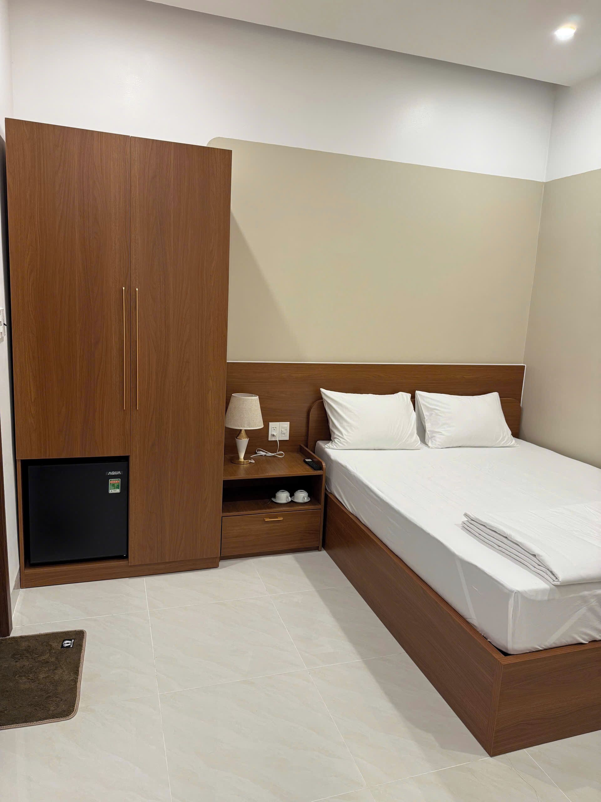 A hotel room with a bed, a wardrobe, a nightstand with a lamp, and a mini-fridge.