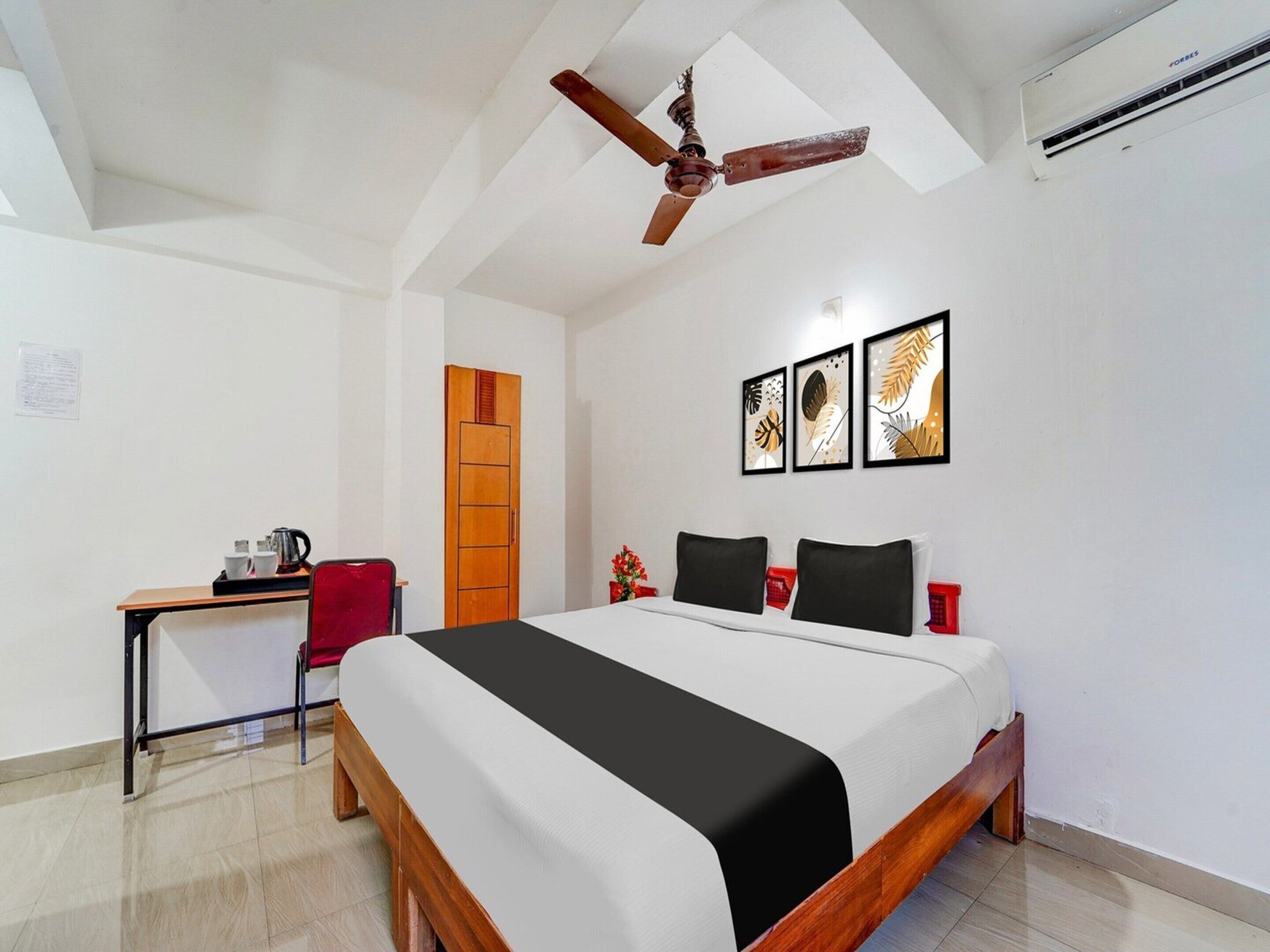 A hotel room with a bed, a desk, a chair, a ceiling fan, and three framed artworks on the wall.