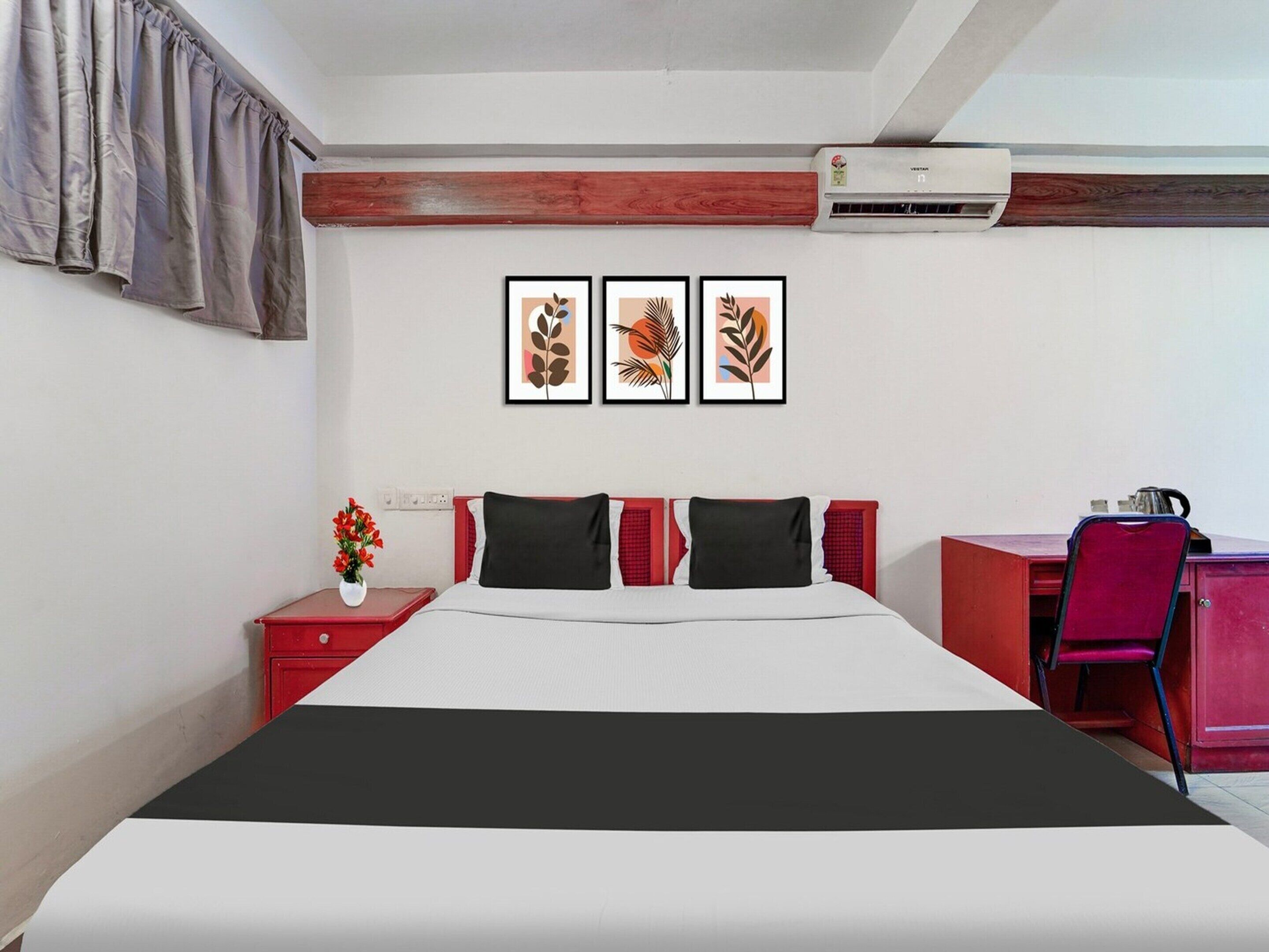 A hotel room with a bed, a desk, a chair, a red cabinet, and three framed artworks on the wall.