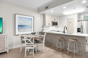A modern kitchen with white cabinetry, a dining area with white chairs, and a bar stool. - Beach Resort 210 - Scenic Gulf Drive! Massive Lagoon Pool! Step to the Beach! (Miramar Beach)