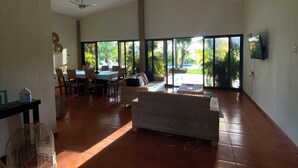 A spacious living area with a large window, a ceiling fan, and a dining area with chairs and a table. - Villa Privada con Piscina y Vista al Estero (El Zapote)