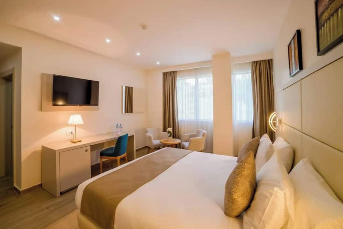 Habitación de hotel con cama, escritorio, sillas, televisor y ventana con cortinas.