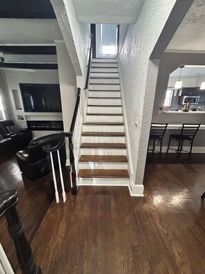 Un escalier menant à une chambre à coucher située en mezzanine, avec vue sur le ciel, un coin repas avec une cuisine en arrière-plan, et un salon équipé d’un téléviseur et de sièges.