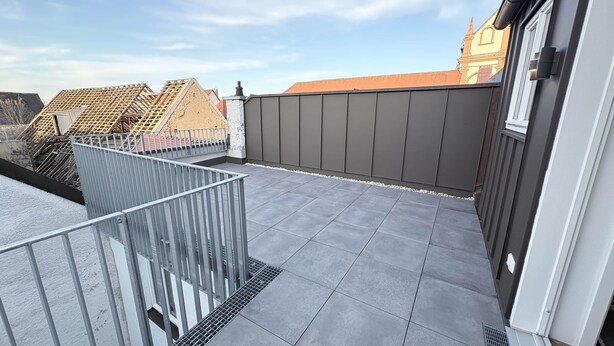 A rooftop terrace with a grey tiled floor, metal railing, and a view of buildings in the background. - 247 Reside: Apartment SZ3 | Center | Netflix (Straubing)