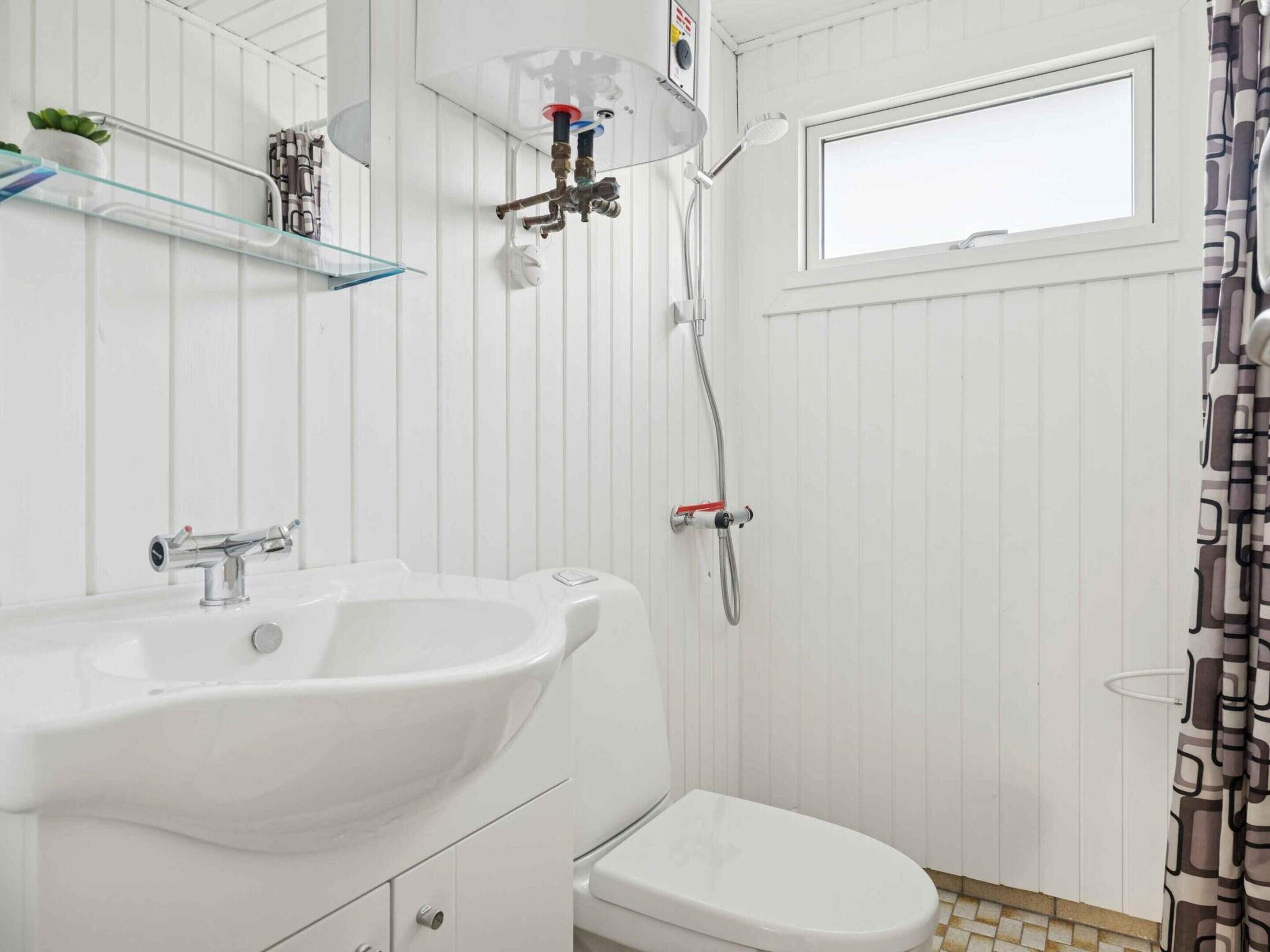 A bathroom with a white sink, toilet, and shower. There is a window above the toilet and a showerhead mounted on the wall.
