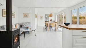 A modern interior with a wood-burning stove, white walls, a dining area, and large windows. - 4 Star Holiday Home in Vaeggerlose (Vaeggerlose)