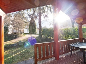 Una terraza de madera con un banco y una mesa, con vistas a un jardín con árboles y una casa al fondo.