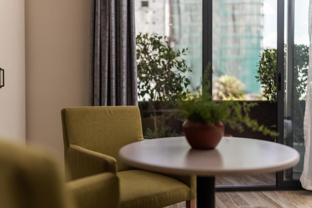 Una habitació amb una butaca verda, una taula rodona petita amb una planta en un test, i una finestra gran amb vistes a edificis i vegetació.