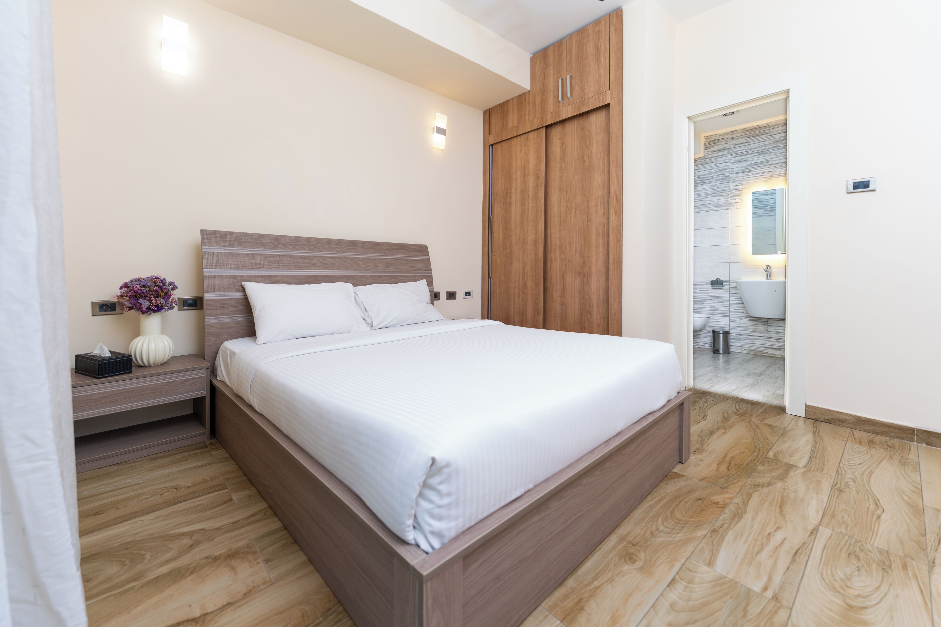 Un dormitorio moderno con estructura de cama de madera, una mesita de noche con un florero y un baño visible a través de una puerta abierta.